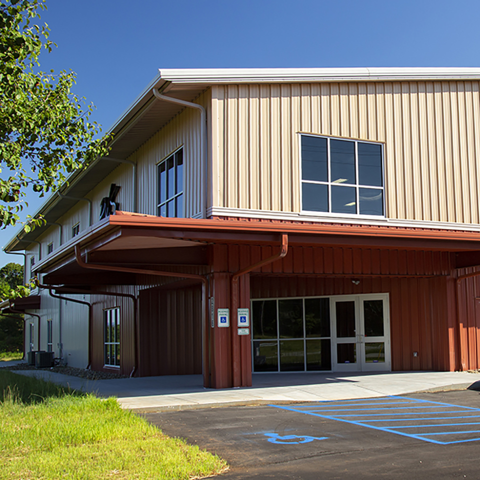 A low-angle exterior photo of a modern, two-story building with a metal facade. The top floor has beige vertical siding and large windows, while the bottom floor is covered with dark red siding. A dark red awning extends over the main entrance, which is a set of glass doors. A paved walkway leads to the entrance, with a handicapped parking space logo painted on the ground. A tree and green grass are visible on the left side of the image under a clear blue sky.